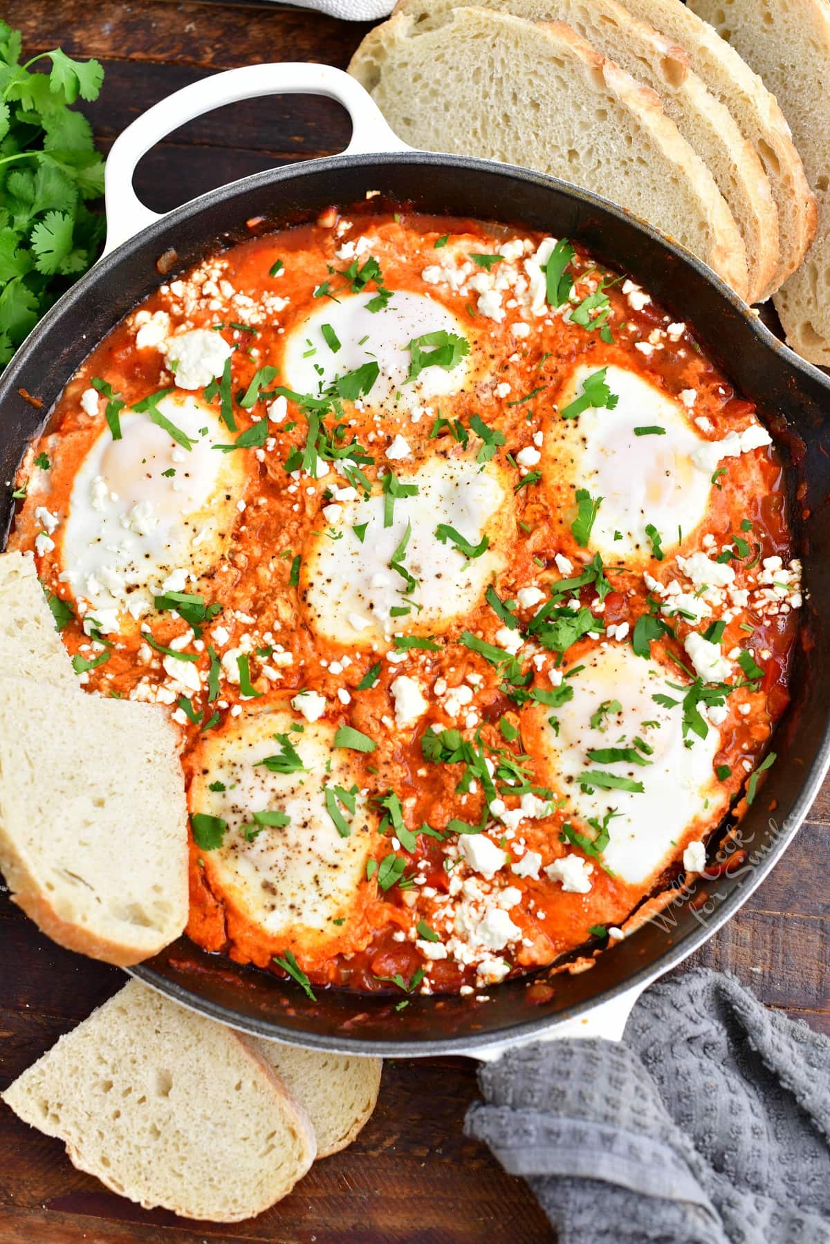 A few slices of bread are placed next to a black skillet filled with shakshuka.