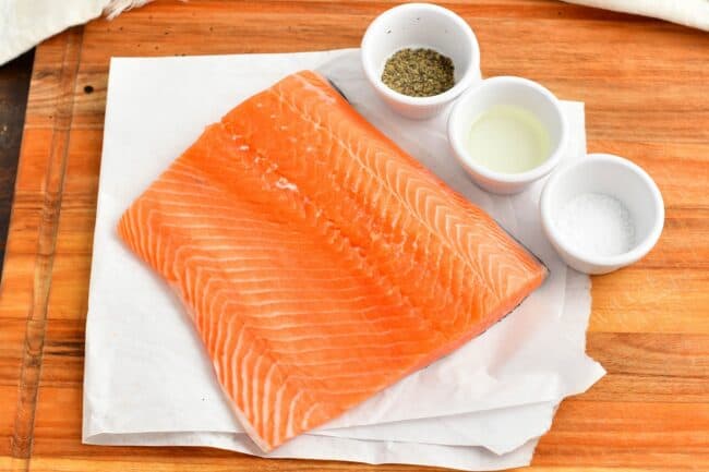 Salmon is placed on a white paper next to cups of seasoning. 