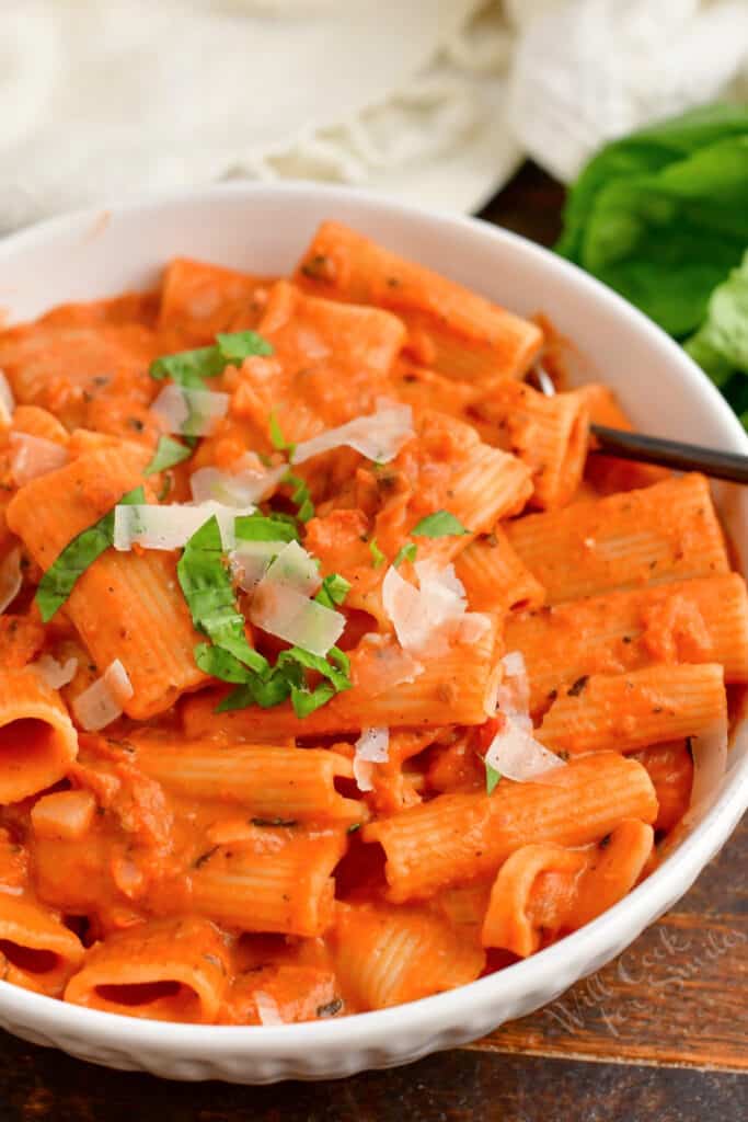 A white bowl filled with pasta is topped with basil and parmesan cheese. 