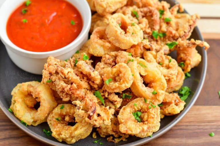 A plate of crispy fried calamari garnished with chopped parsley, served with a small bowl of red marinara sauce on the side.