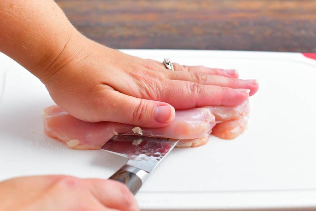 A hand is pressing raw chicken down while a knife is slicing through the middle of the breast.