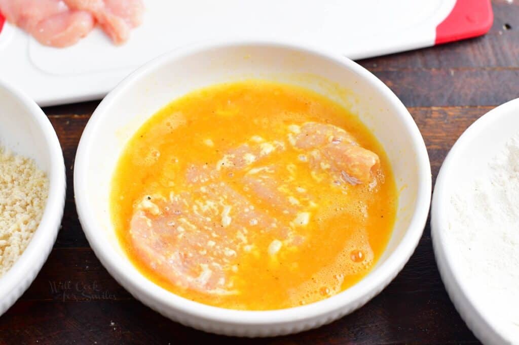 The chicken is being dipped into a white bowl filled with egg mixture.