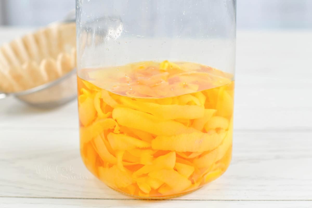 Lemon peels and vodka are combined in a glass jar that's placed on a white countertop.