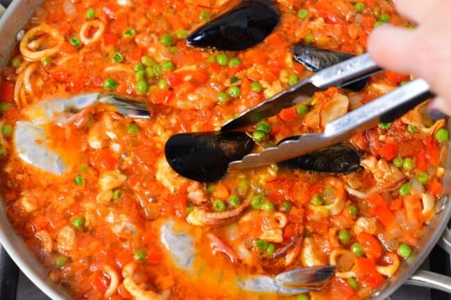 Muscles are being added to the paella with tongs.