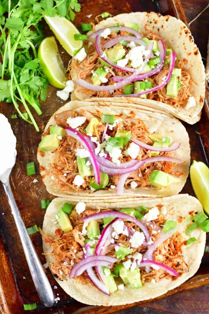 three shredded chicken tacos with toppings on a sheet pan with sour cream and greens next to them