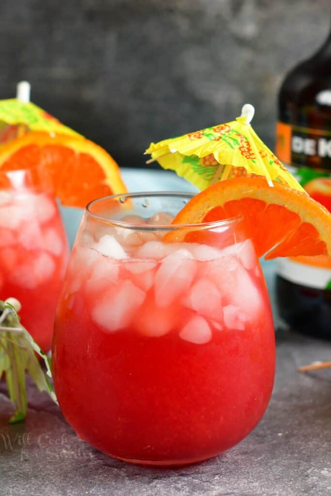 A glass of a vibrant red cocktail is garnished with an orange wedge and an umbrella. 