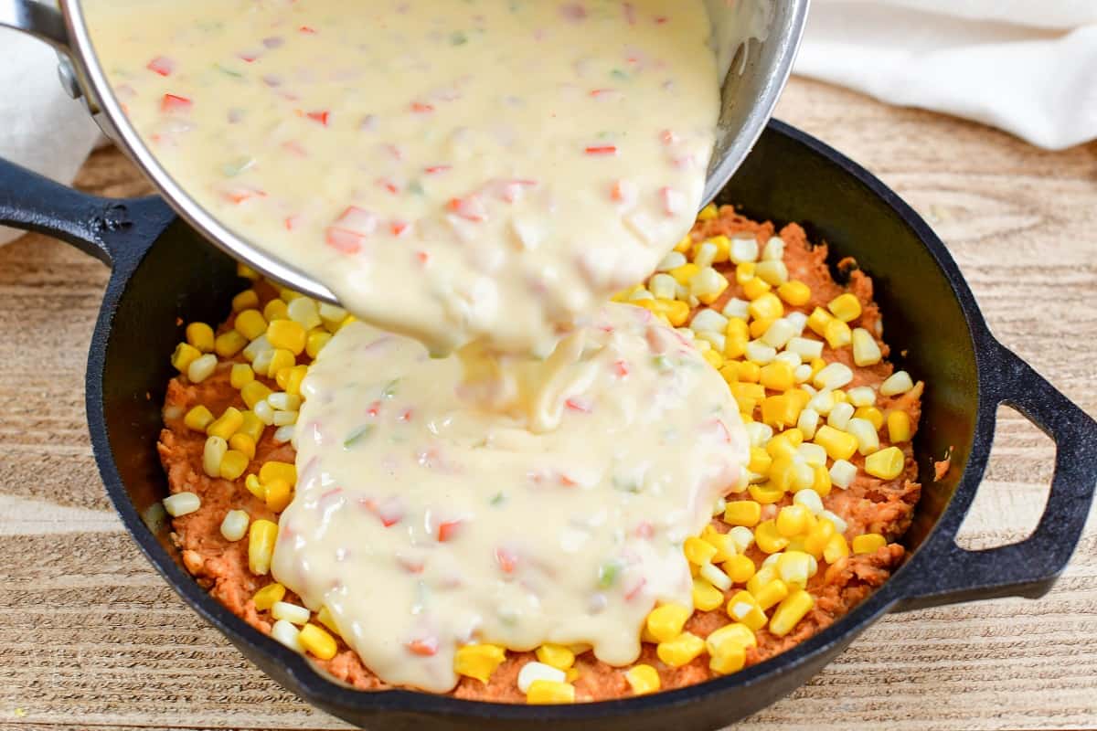 pouring queso mixture over refried bean and corn