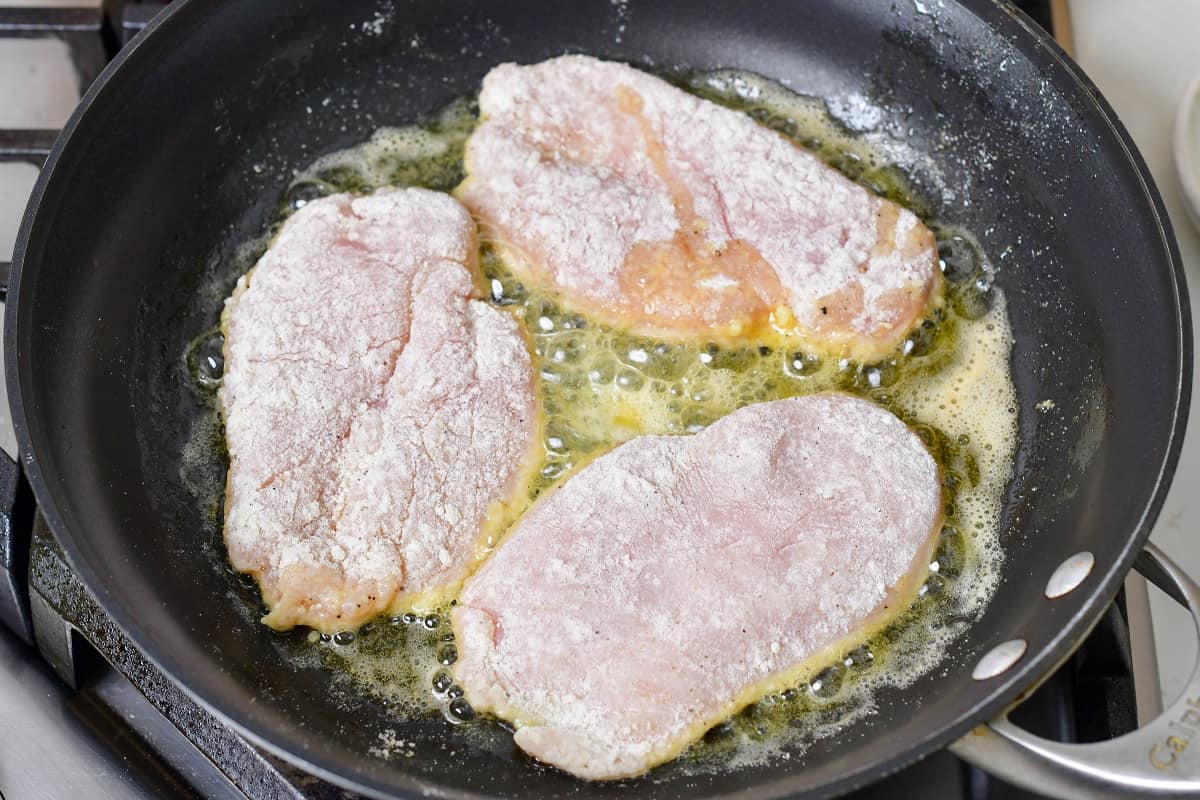 three dredged chicken cutlets searing in a pan