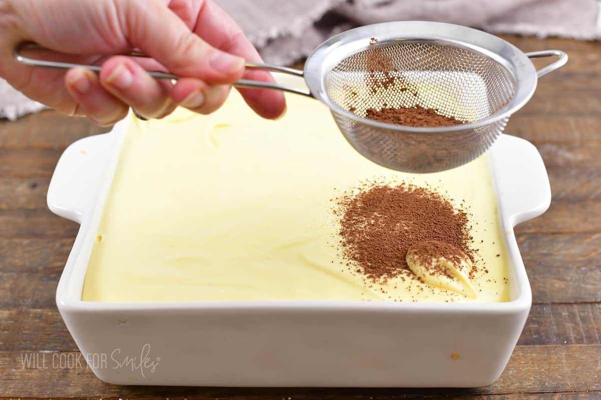 dusting layered tiramisu in a baking with with cocoa.
