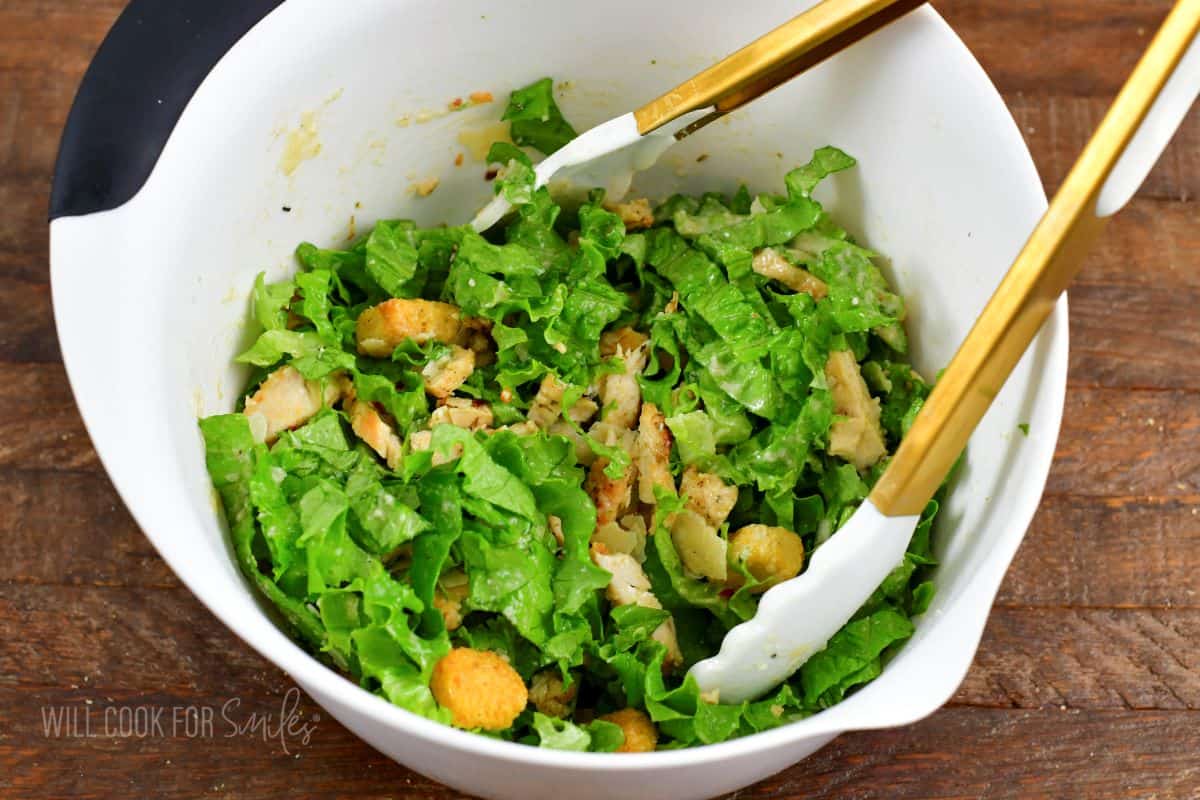tossing chicken Caesar salad in a bowl with tongs.