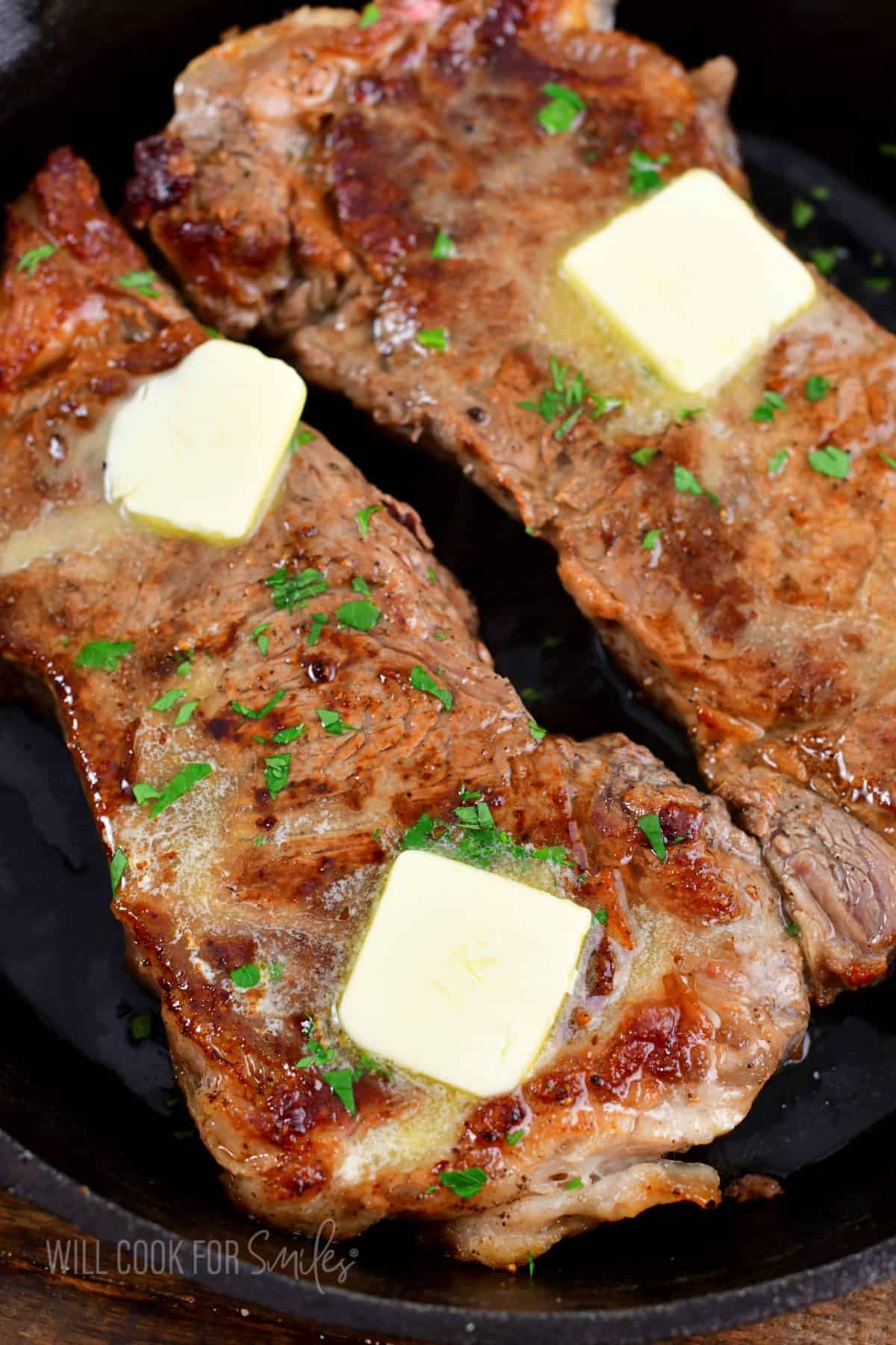 two cooked steaks topped with butter and parsley in a skillet.