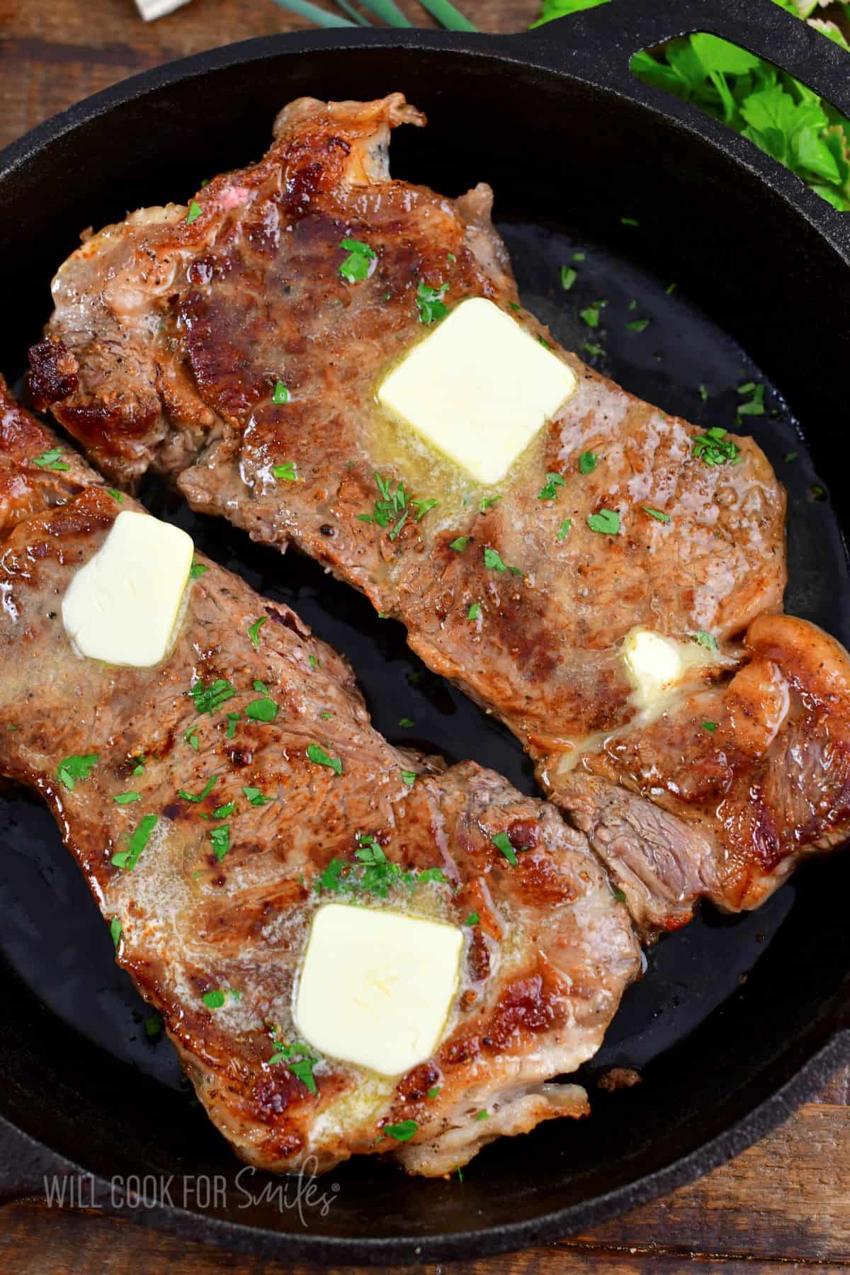 butter slices melting on top of two cooked steaks in a cast iron skillet.