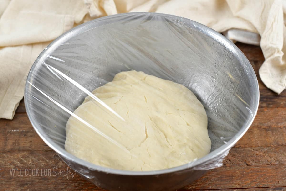 Dough for dinner rolls in a meatal bowl with plastic cover.
