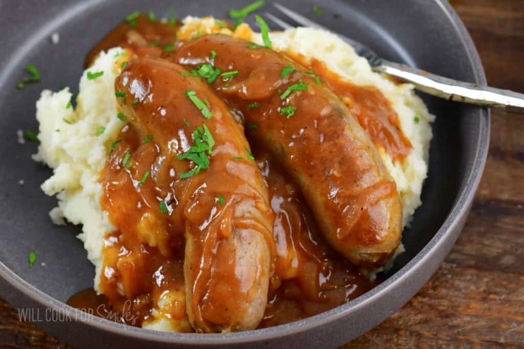 sausages in onion gravy over mashed potatoes in a plate.