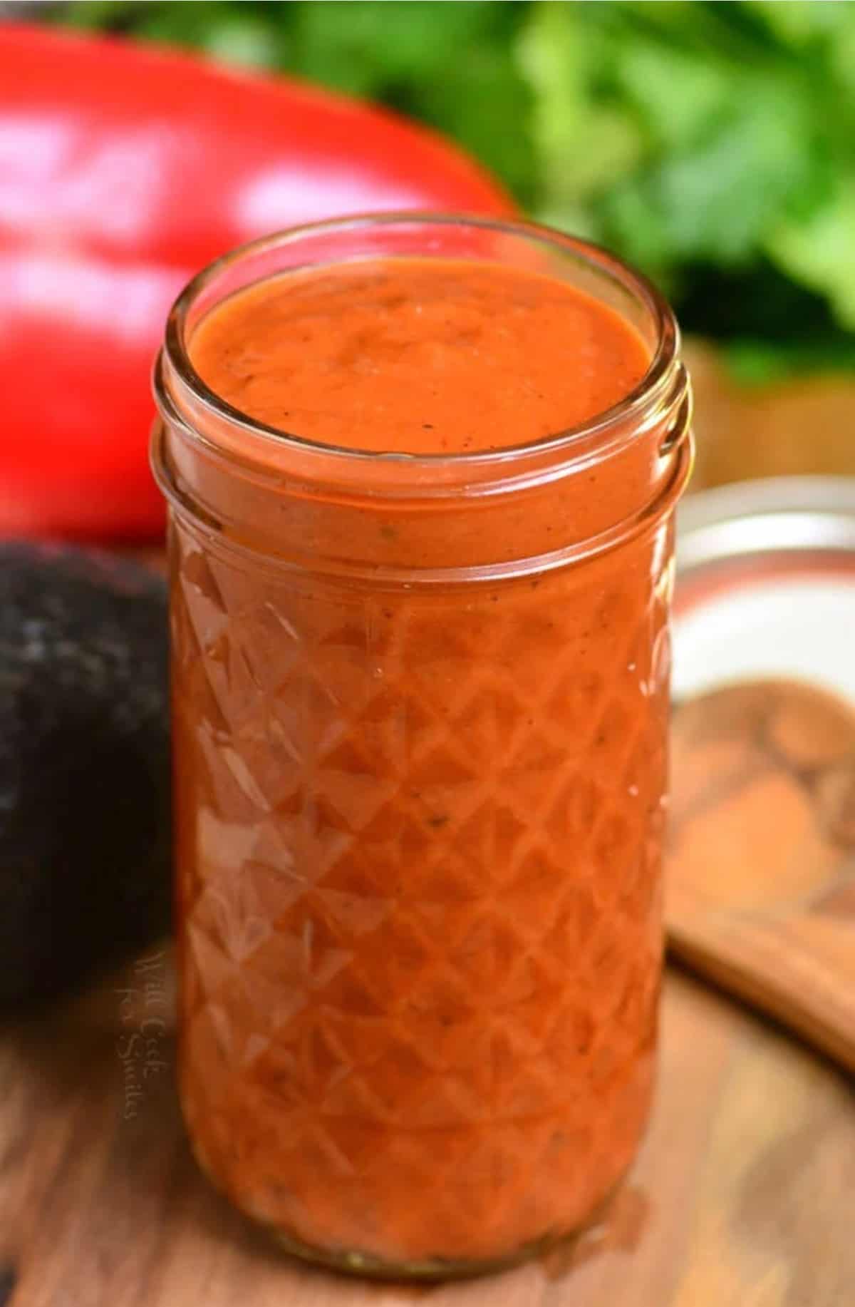 tall glass jar full of enchilada sauce on wooden surface with veggies around.
