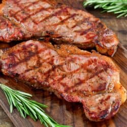 two grilled steaks on the wooden cutting board with rosemary around.