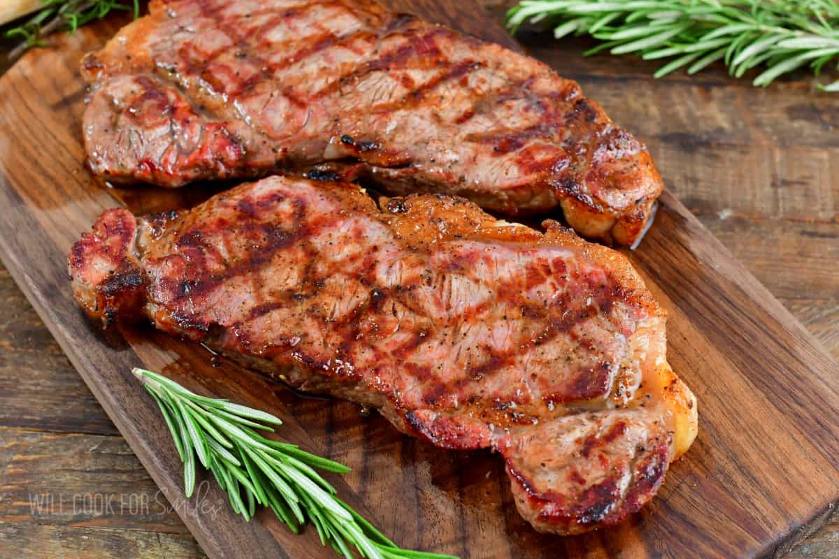 two grilled steaks on the wooden cutting board with rosemary around.