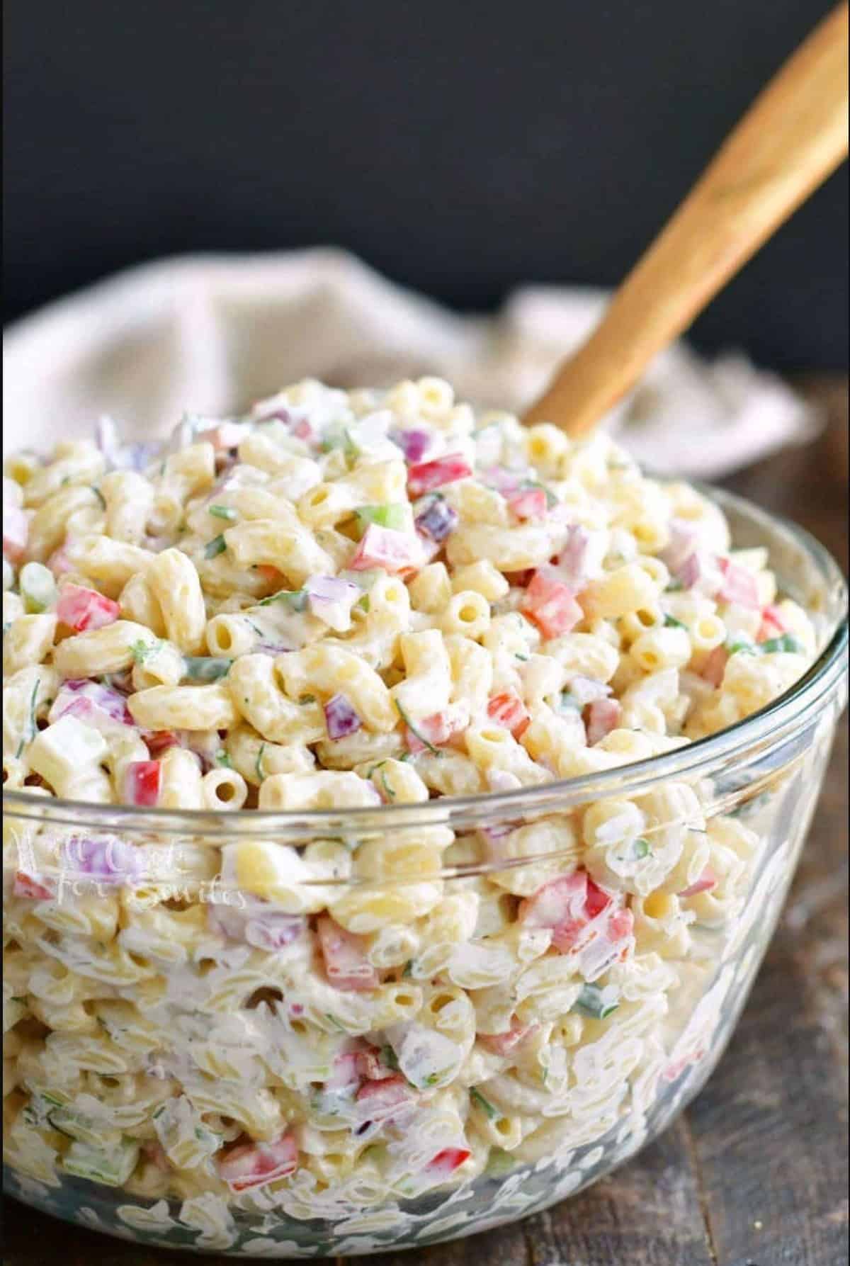 a large glass bowl filled with colorful creamy macaroni salad and a wooden spoon.