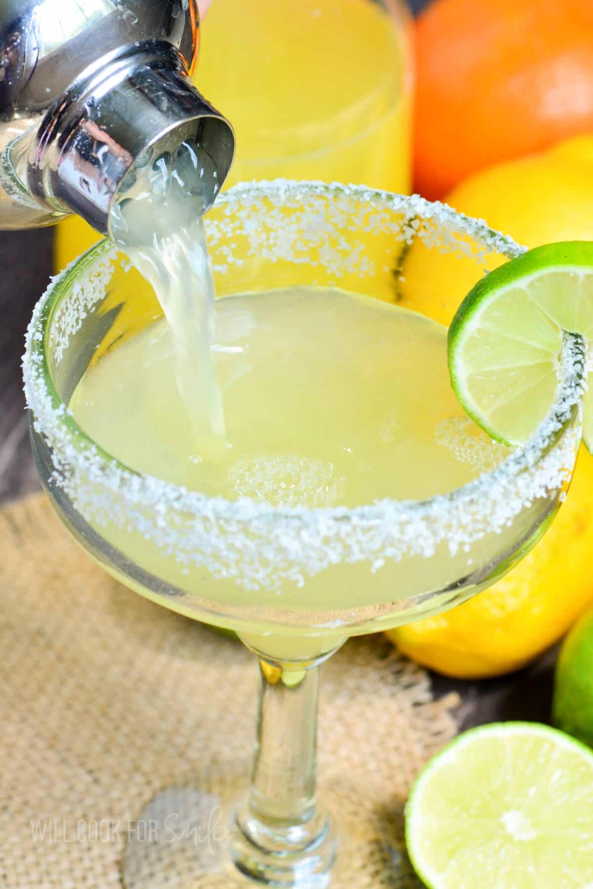 pouring light colored margarita cocktail into glass with salted rim.