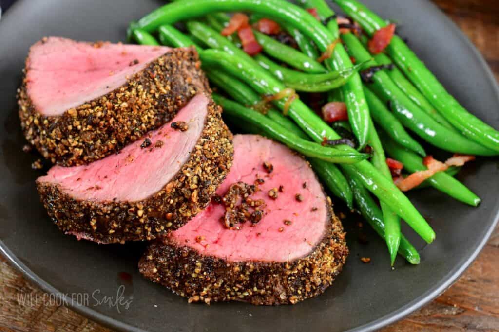 A plate with sliced, seasoned, medium-rare beef tenderloin accompanied by green beans sautéed with bacon pieces. The dish is arranged neatly with three beef slices showing a pink center and pepper crust. The green beans are vibrant and glistening.