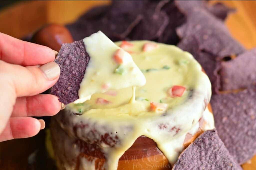 hand holding a blue tortilla chip with queso dip on it above a crock filled with queso.
