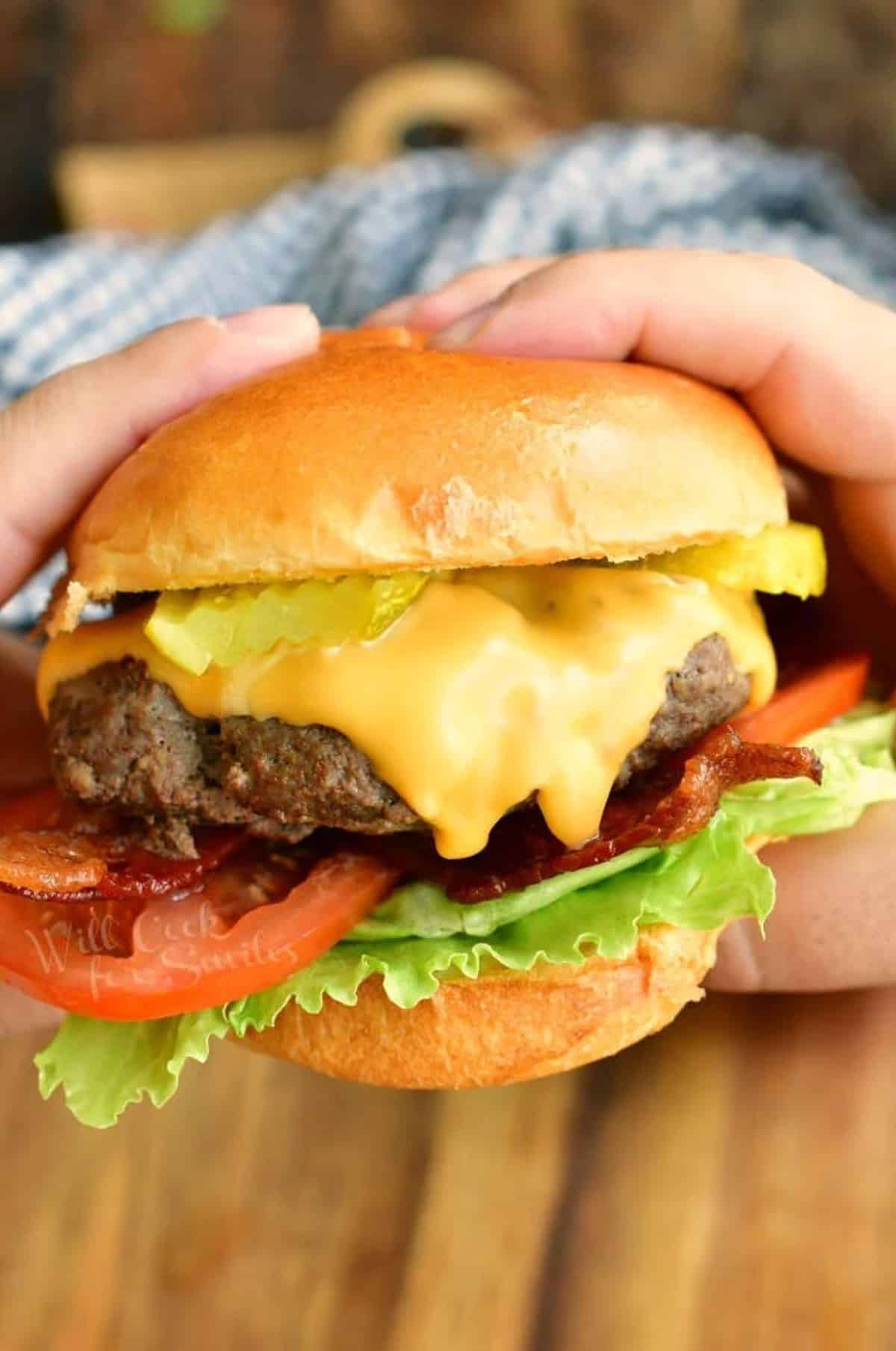 holding a cheeseburger with tomatoes lettuce and pickles with two hands.