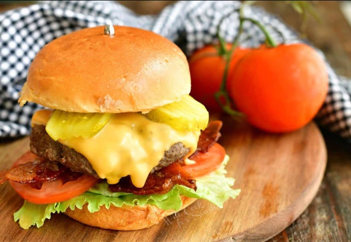 a cheeseburger on a wooden board with tomatoes on a vine and a checkered towel.