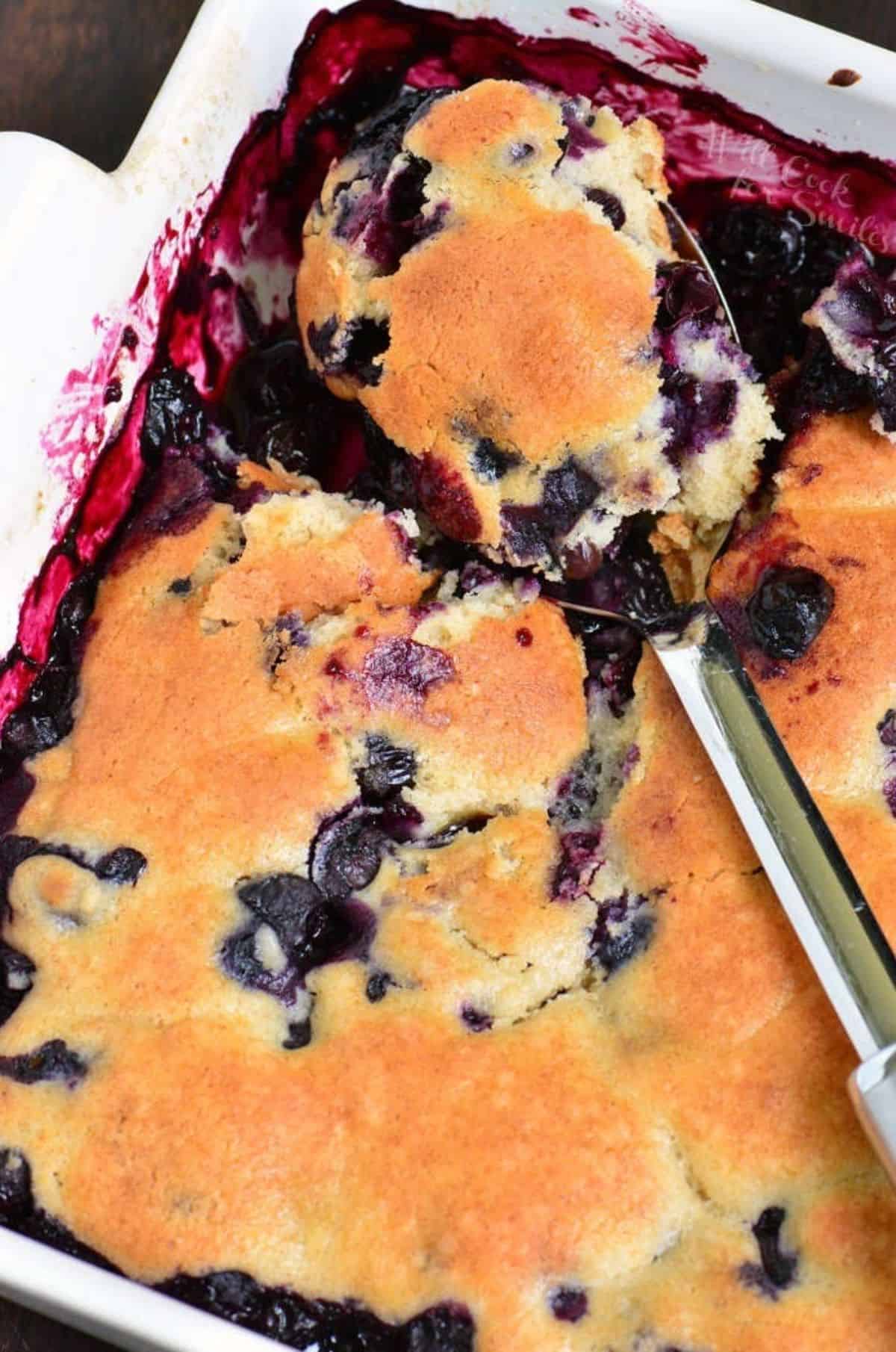 blueberry cobbler in a white baking dish with ladleful on top.