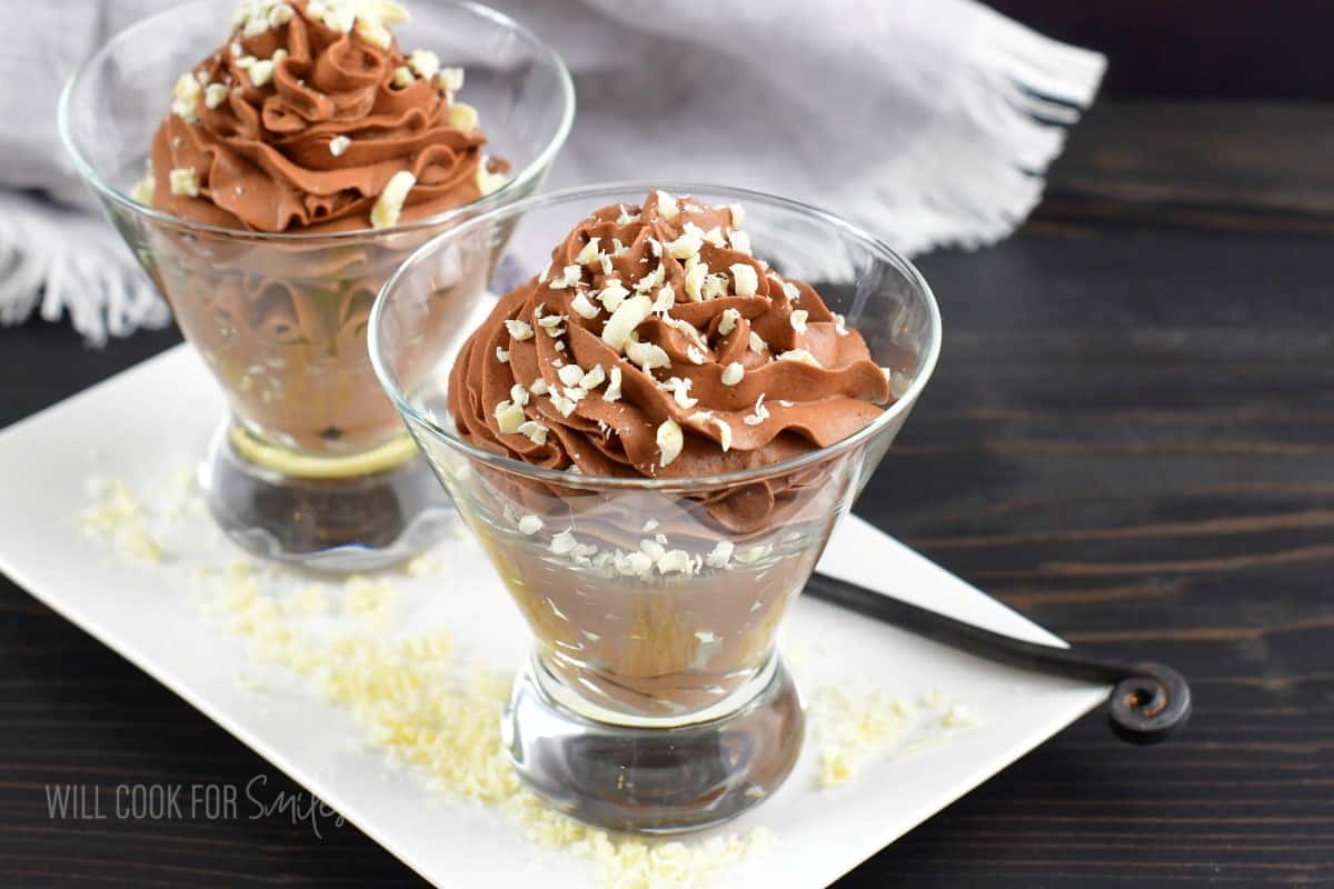 two clear glasses filled with chocolate mousse and topped with white chocolate.