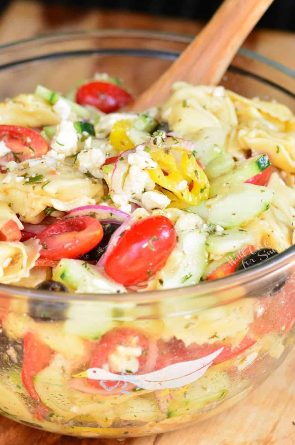 mixed Greek tortellini salad in a glass bowl with a wooden spoon.