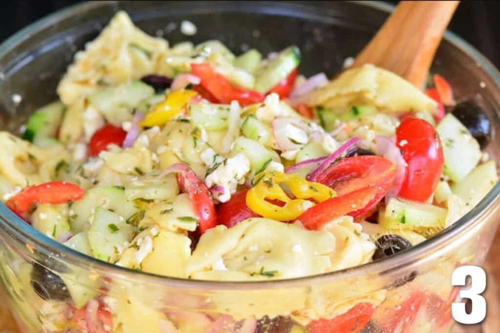 mixing Greek tortellini salad in a glass bowl.