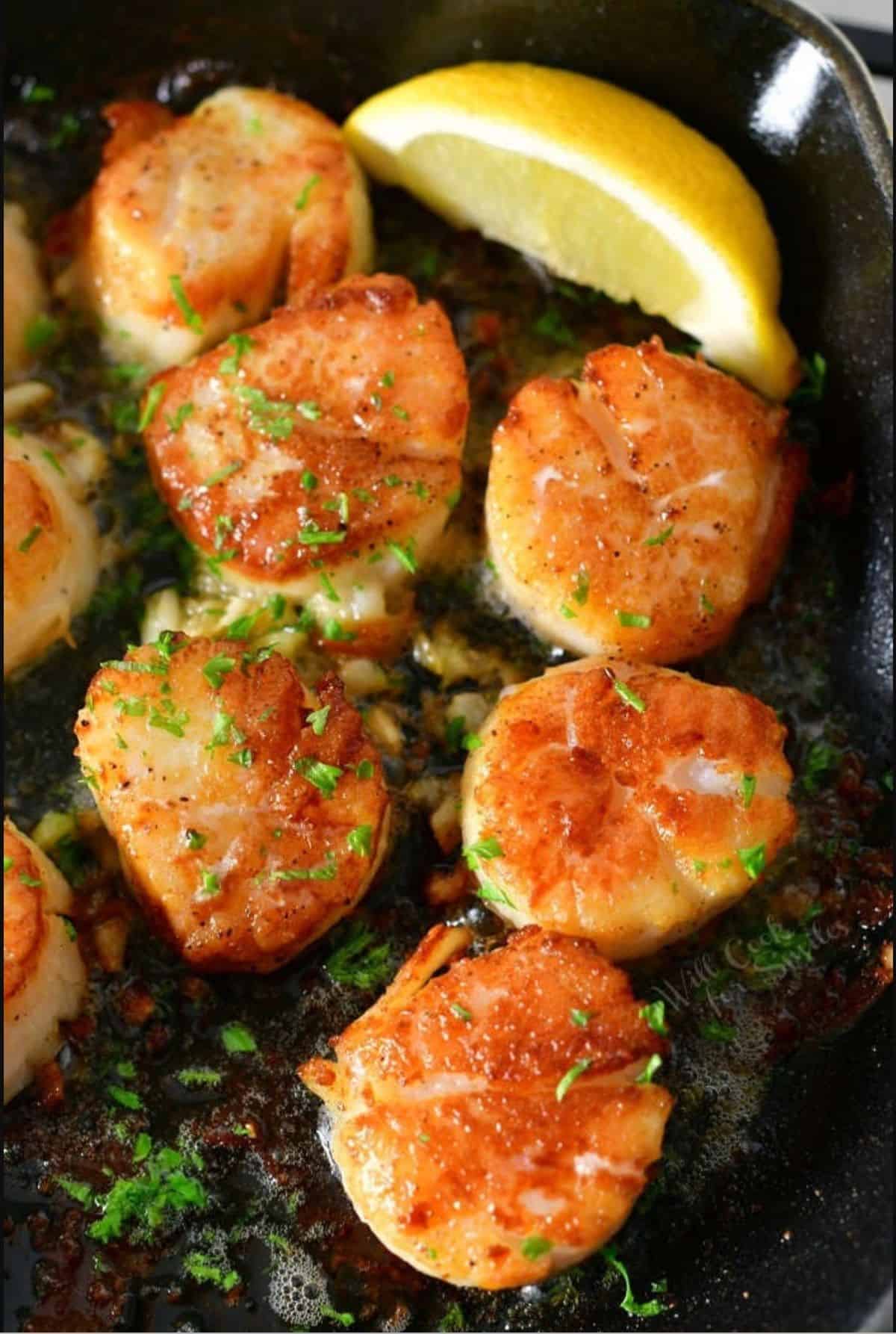 several seared scallops in a skillet with parsley sprinkled on top and a wedge of lemon.