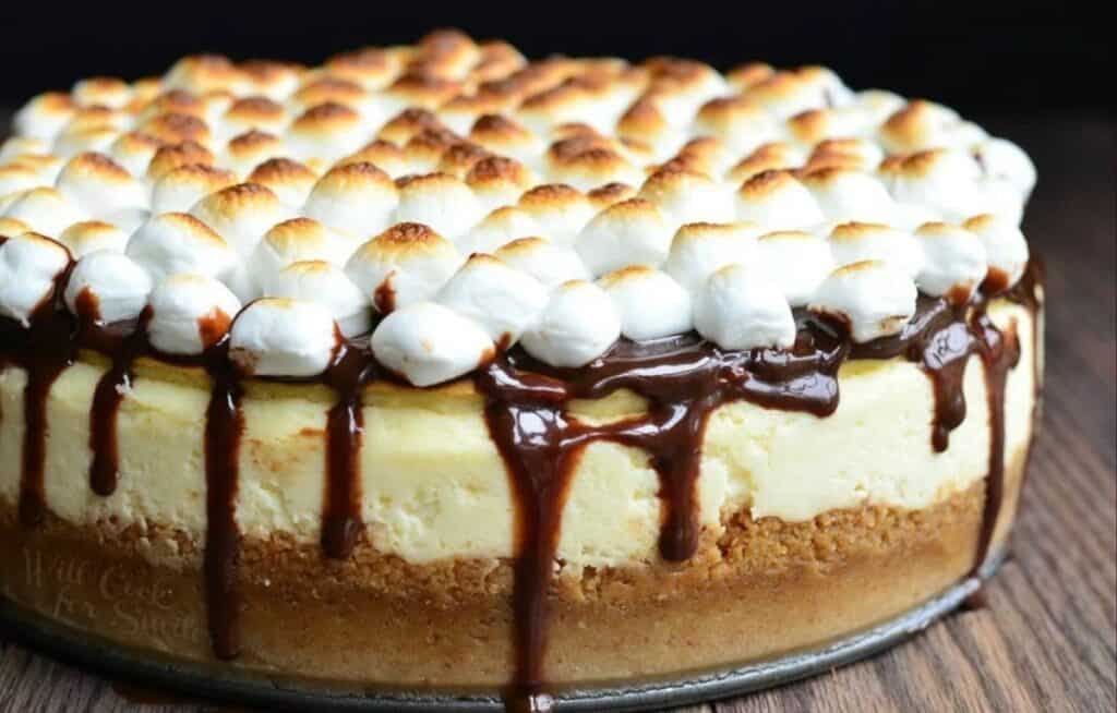 A cheesecake topped with toasted marshmallows and drizzled chocolate sits on a wooden surface. The layers include a graham cracker crust, creamy filling, and a rich chocolate drizzle.
