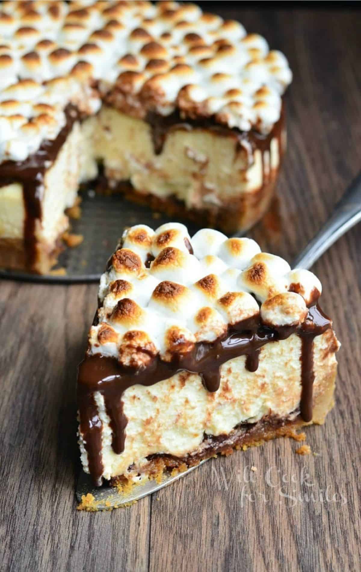 cut out slice of smores cheesecake next to a while cheesecake on wooden board.