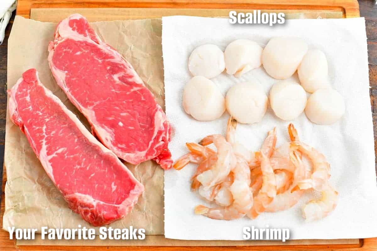 two steaks, several scallops, and several shrimp on the cutting board.