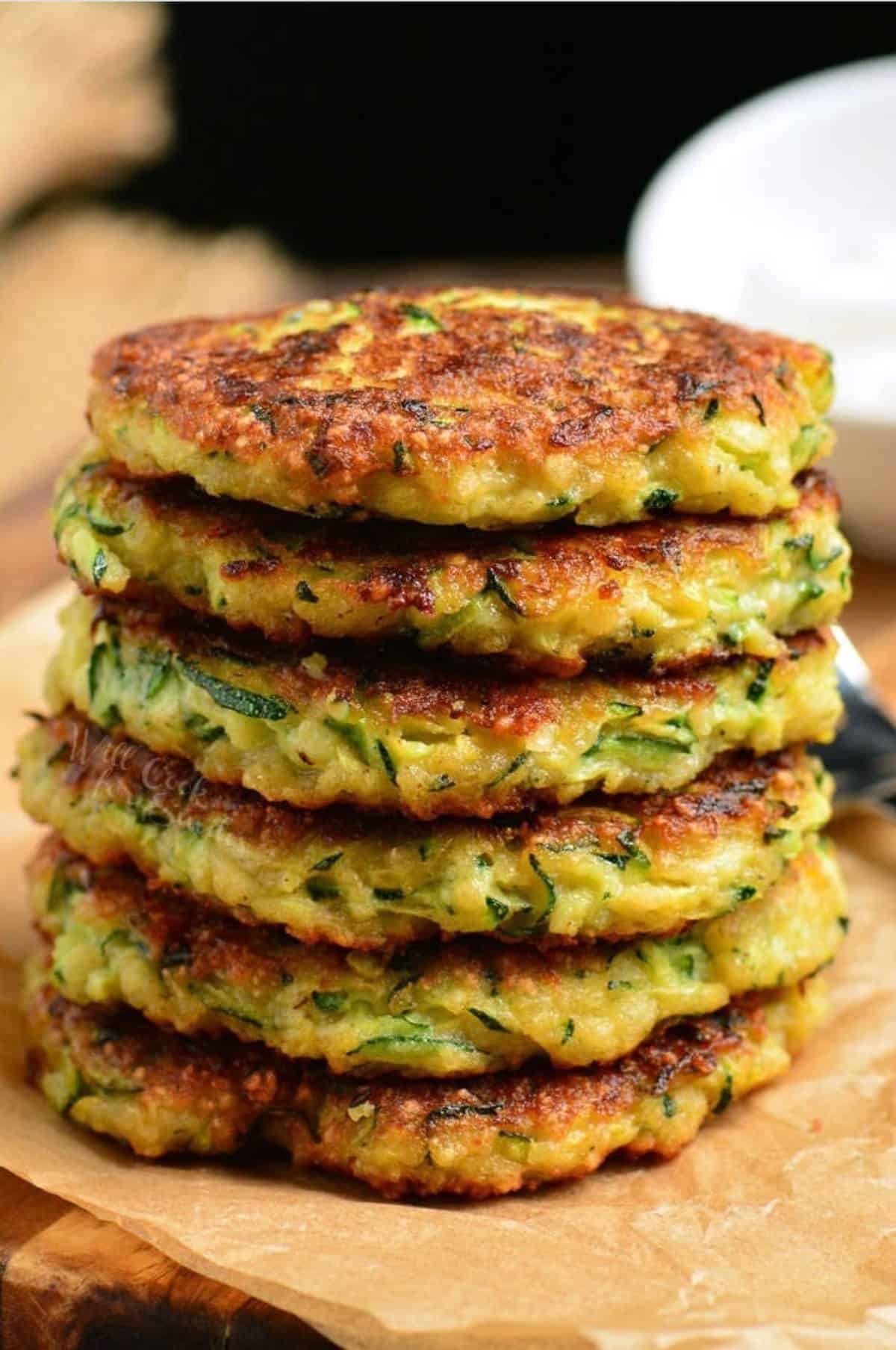 six zucchini fritters stacked on top of each other on parchment paper.