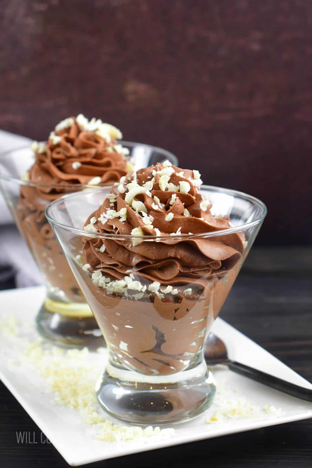 two glasses filled with chocolate mousse topped with shaved white chocolate on white plate.