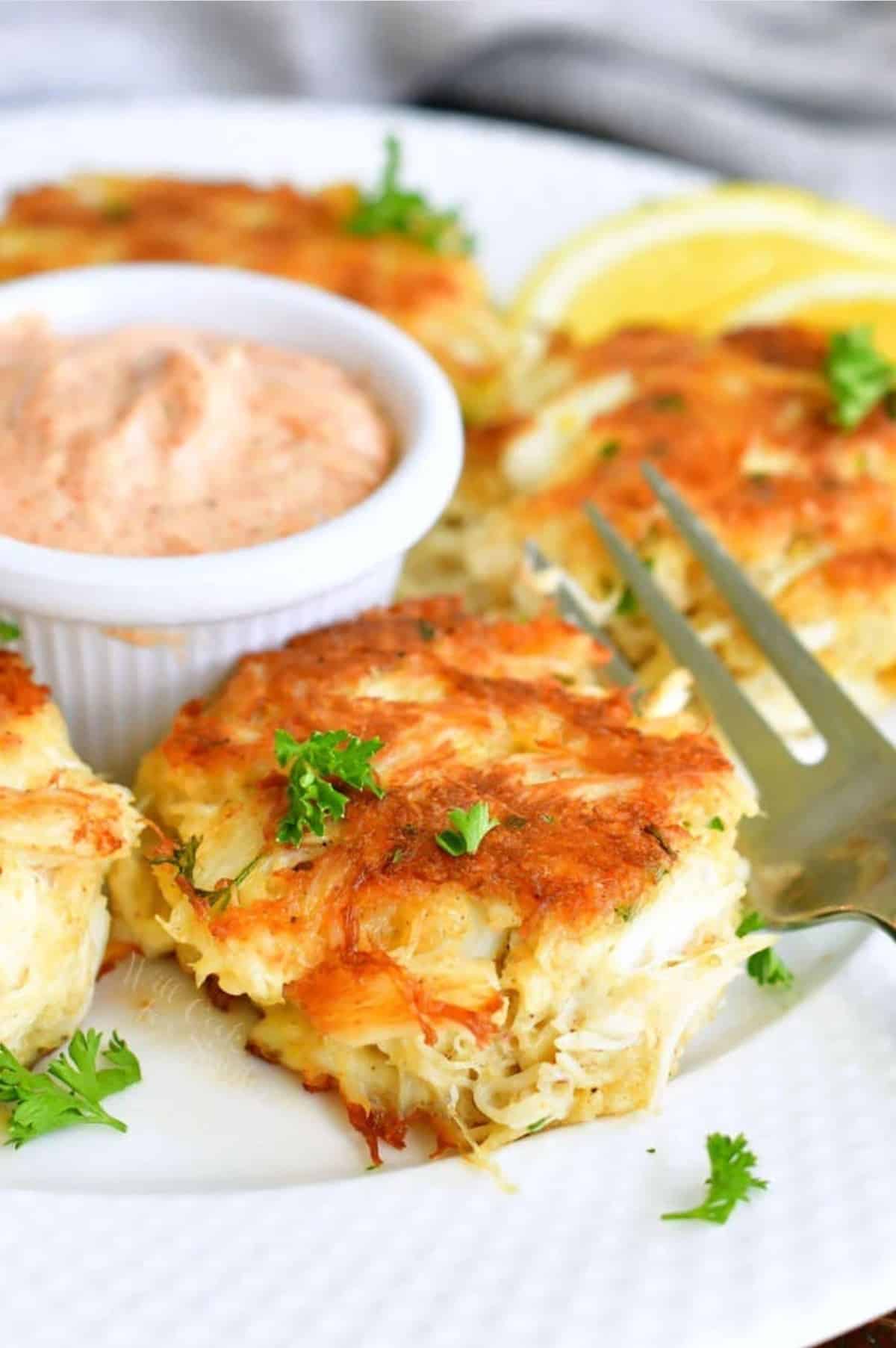 crab cake on a white plate with a fork and remoulade sauce in white cup.