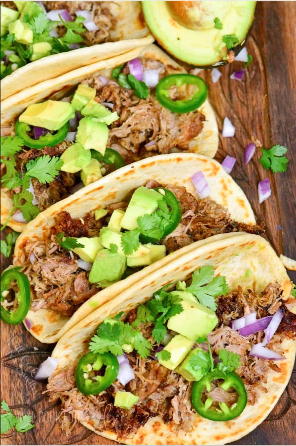 four pork carnitas tacos laid side by side with toppings like avocado and jalapenos.