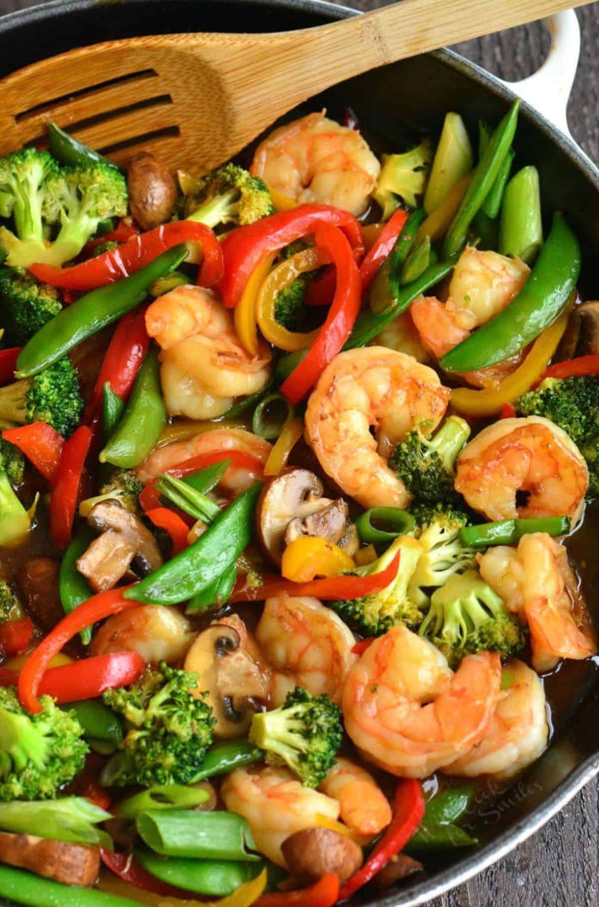 shrimp and lots of vegetables in sauce in a skillet with a wooden spoon.
