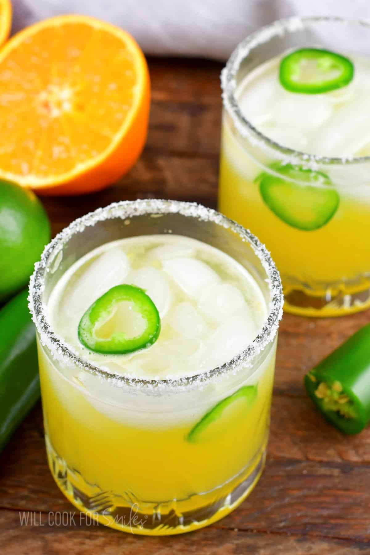two glasses with yellow spicy margarita, salted rim, and sliced jalapenos in it.