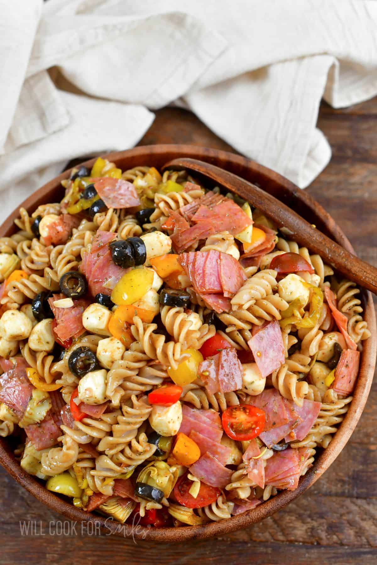 Italian antipasto pasta salad in a wooden bowl with a wooden spoon and a towel.
