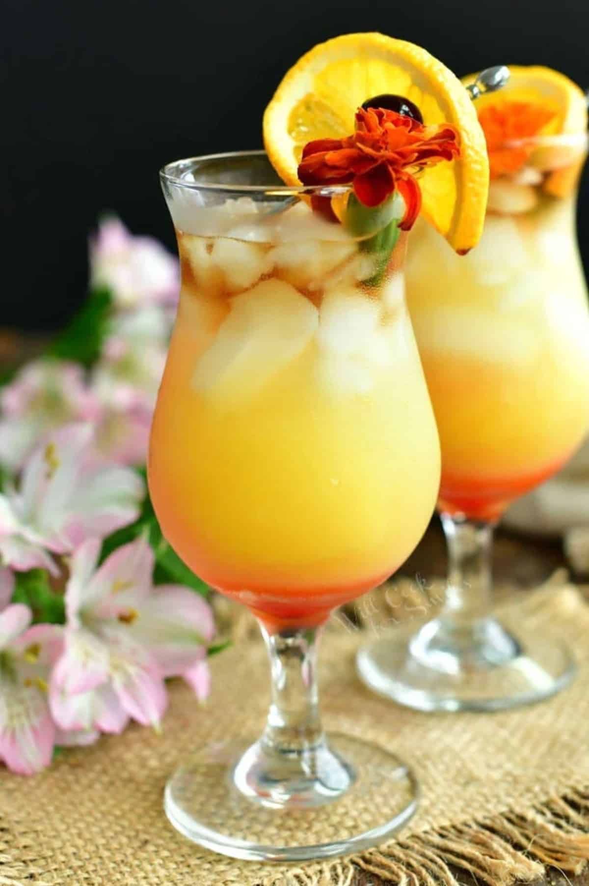 layered cocktail pink on the bottom, orange in the middle, and dark clear on top garnished with a flower and orange slide.