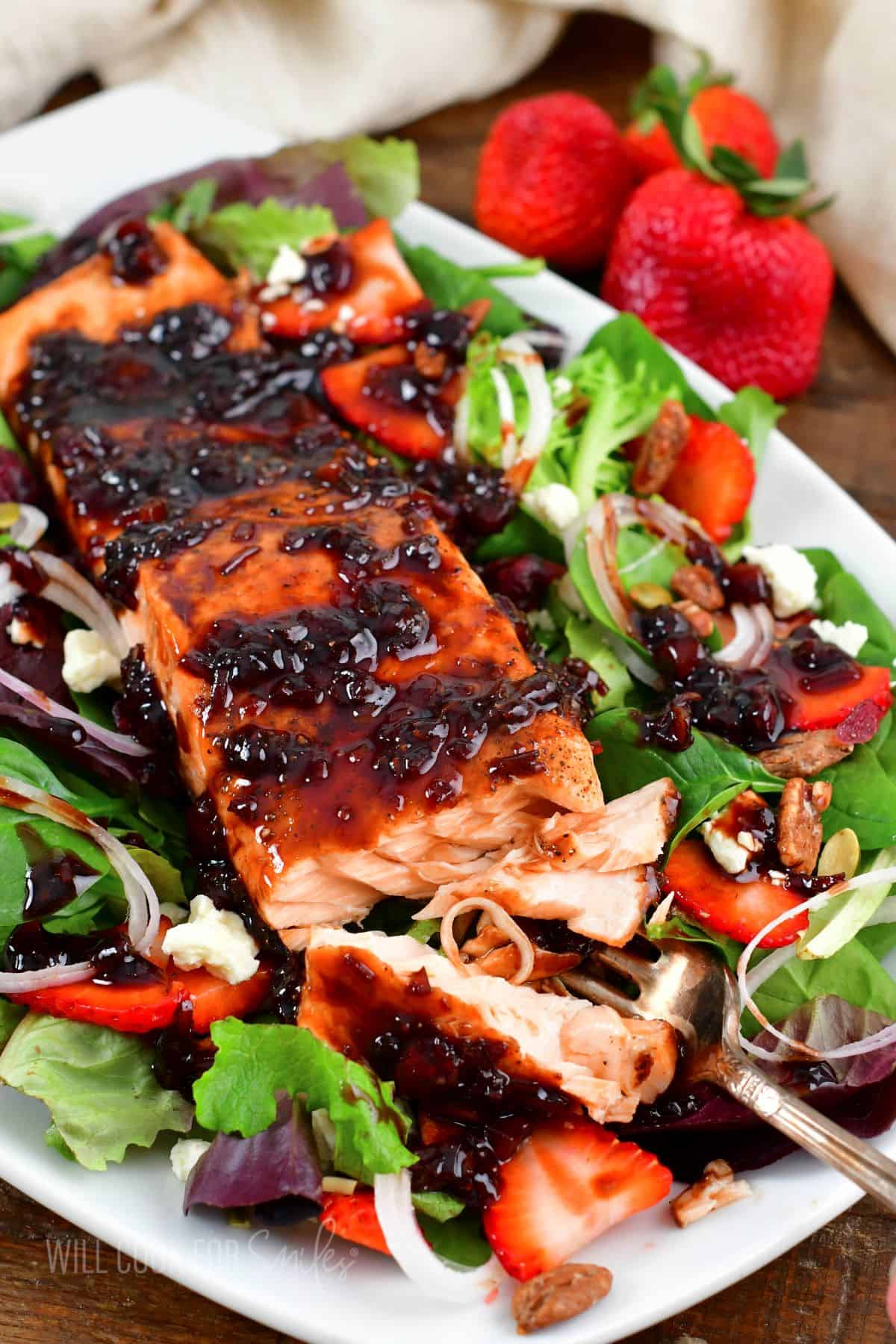 flaking salmon with a fork that's on top of a bright salad with strawberries.