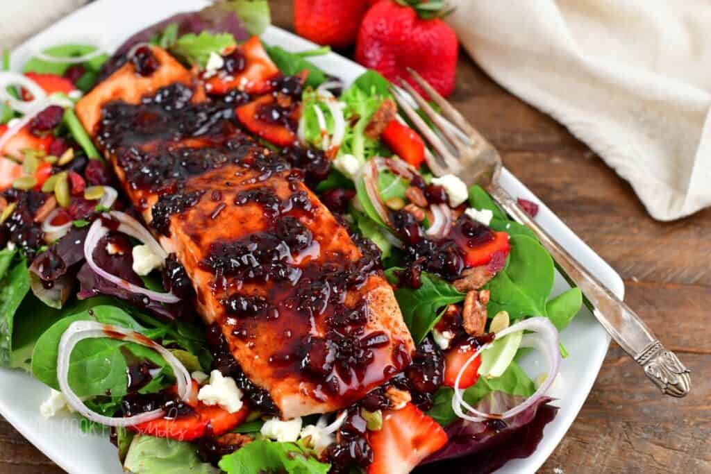 A plate of salad with mixed greens, strawberries, pecans, onions, feta cheese, and a large piece of salmon glazed with a dark sauce. A fork is placed on the plate, and fresh strawberries are in the background.