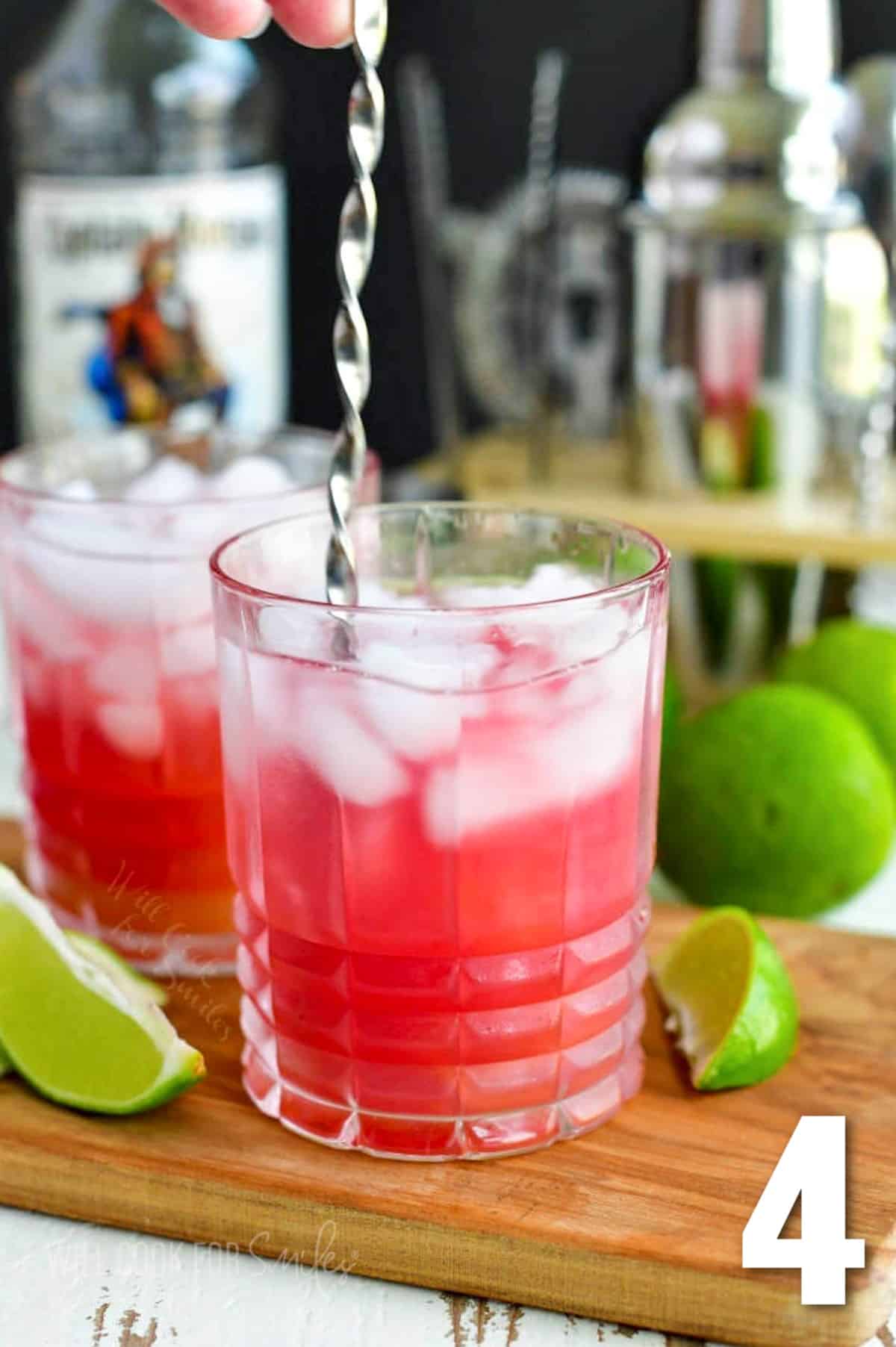 stirring the pink bay breeze cocktail with a bar spoon with limes wedges around.