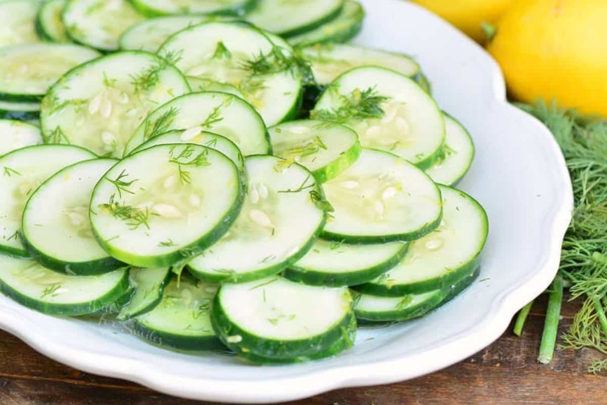 thin sliced cucumbers in cucumber salad in a plate next to dill weed and lemons.