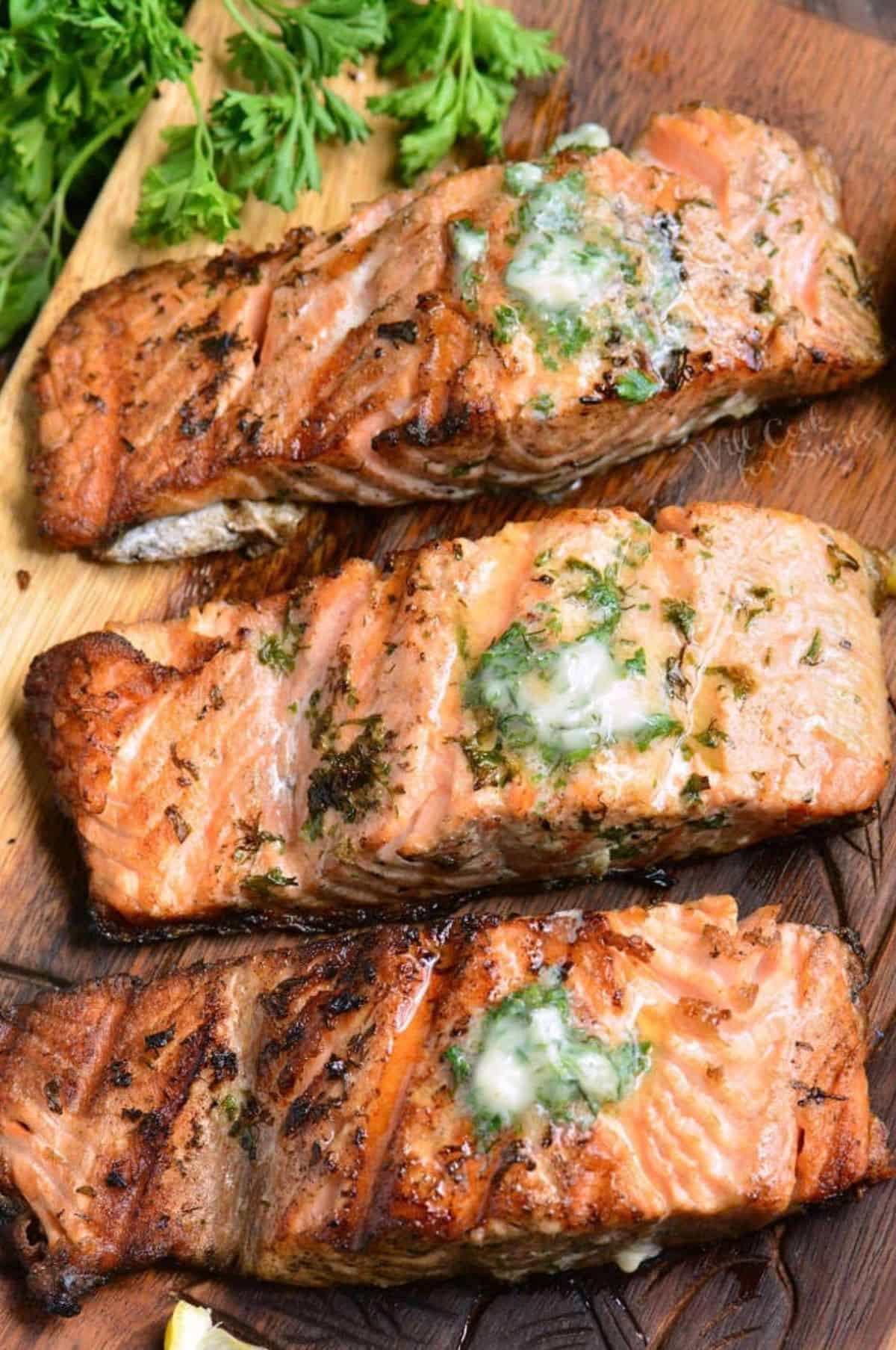 three grilled salmon filets topped with garlic butter on the wooden plate.
