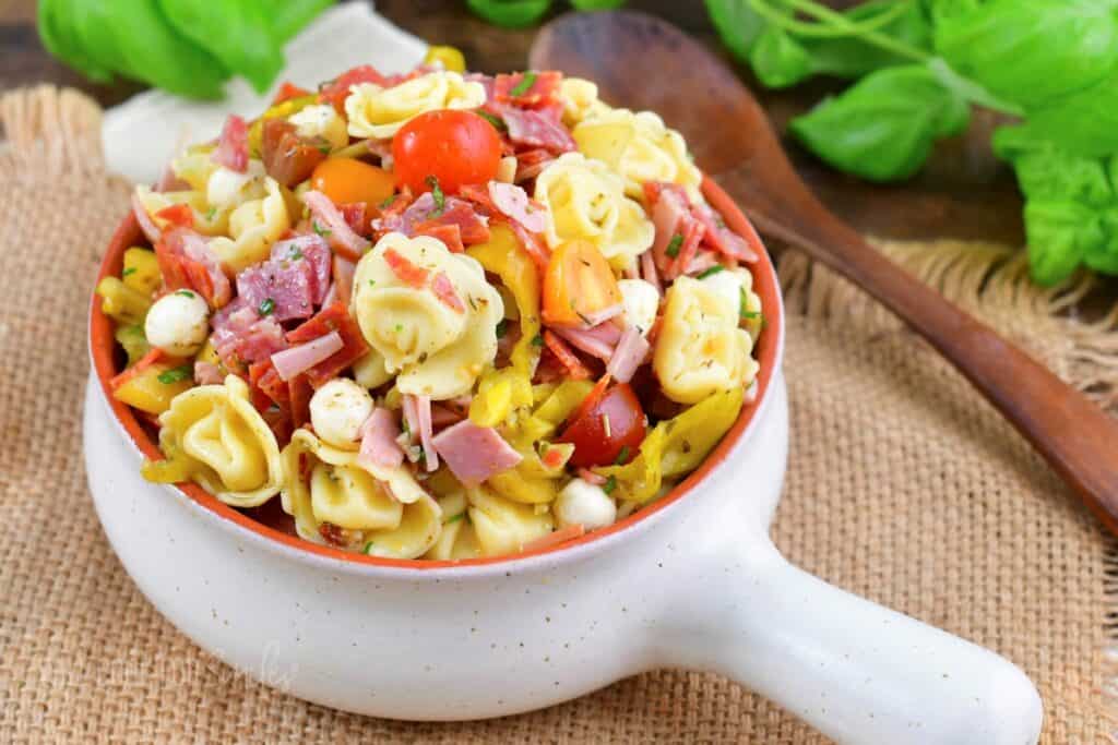 A white bowl filled with a colorful pasta salad, including tortellini, cherry tomatoes, salami slices, cheese, and vegetables, sits on a woven placemat. A wooden spoon and fresh green basil leaves are visible in the background.