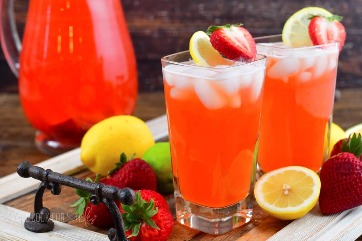 two glasses filled with strawberry lemonade on a tray with fruit around and a pitcher.